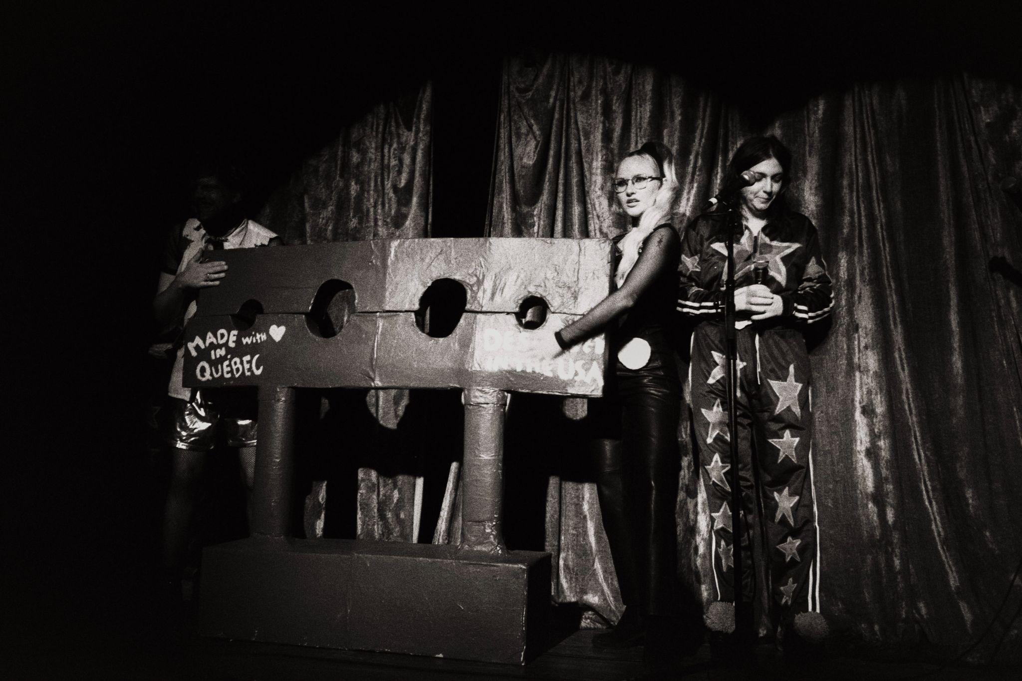 Two spelling bee hosts stand beside contestants in a large cardboard guillotine costume